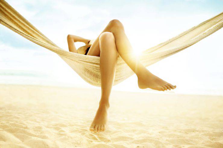 Woman relaxes in a hammock with bare feet.  Her feet and legs are featured in the foreground of the image.  One hand is raised to hold a straw hat on her head, though her face is not visible.  The hammock is hung at the beach with the sun shining from behind it.  The hammock is made of lightweight material, which the light passes through.  The sand is soft and has many footprints in it.  A strip of ocean is visible above the sand, and above that is the sky, which is bright blue and has some white clouds.