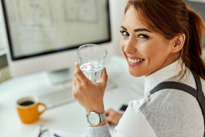 Donna sorride con un bicchiere di acqua in mano e un caffè sulla scrivania