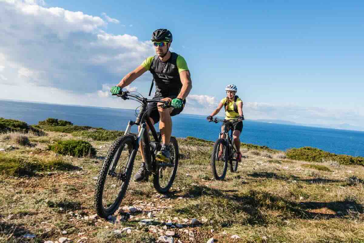 Due ciclisti su strada con vegetazione e mare sullo sfondo