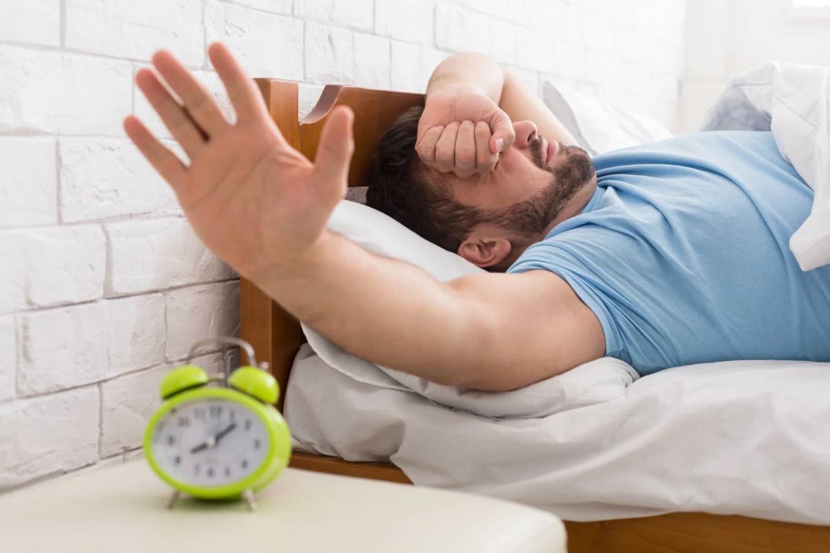 Uomo nel letto che si strofina gli occhi e allunga la mano per spegnere la sveglia