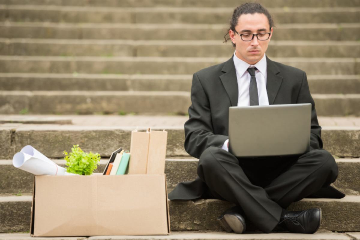 uomo seduto sulle scale per strada con il computer sulle ginocchia e accanto uno scatolone